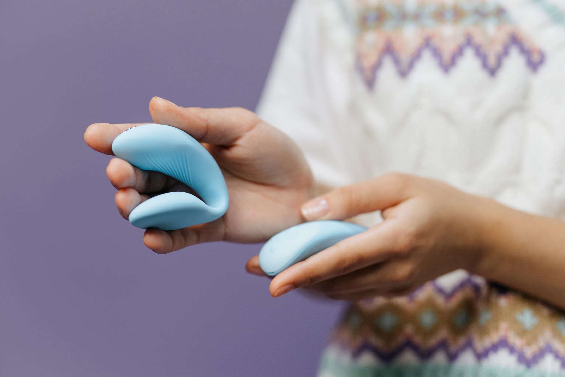 Two hands holding a sleek blue vibrator together against a purple background, symbolizing shared pleasure and intimacy.